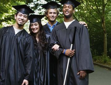  Four smiling students in graduation gowns and caps. The student on the far right is holding a long cane.