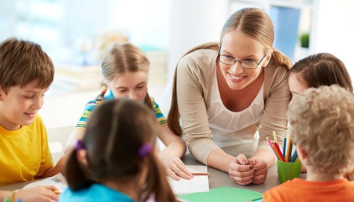 Smiling female teacher leaning over a group of desks with children working