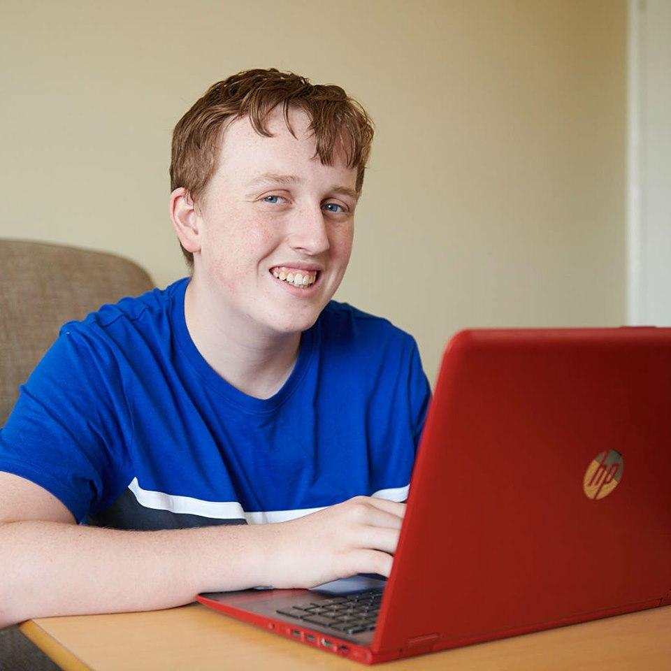 16 year old smiling Charlie wearing a blue t-shirt sat at a table with a red laptop