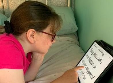 Jessica, a 10 year old girl, lying on her bed, reading on her iPad which is leaning against the wall