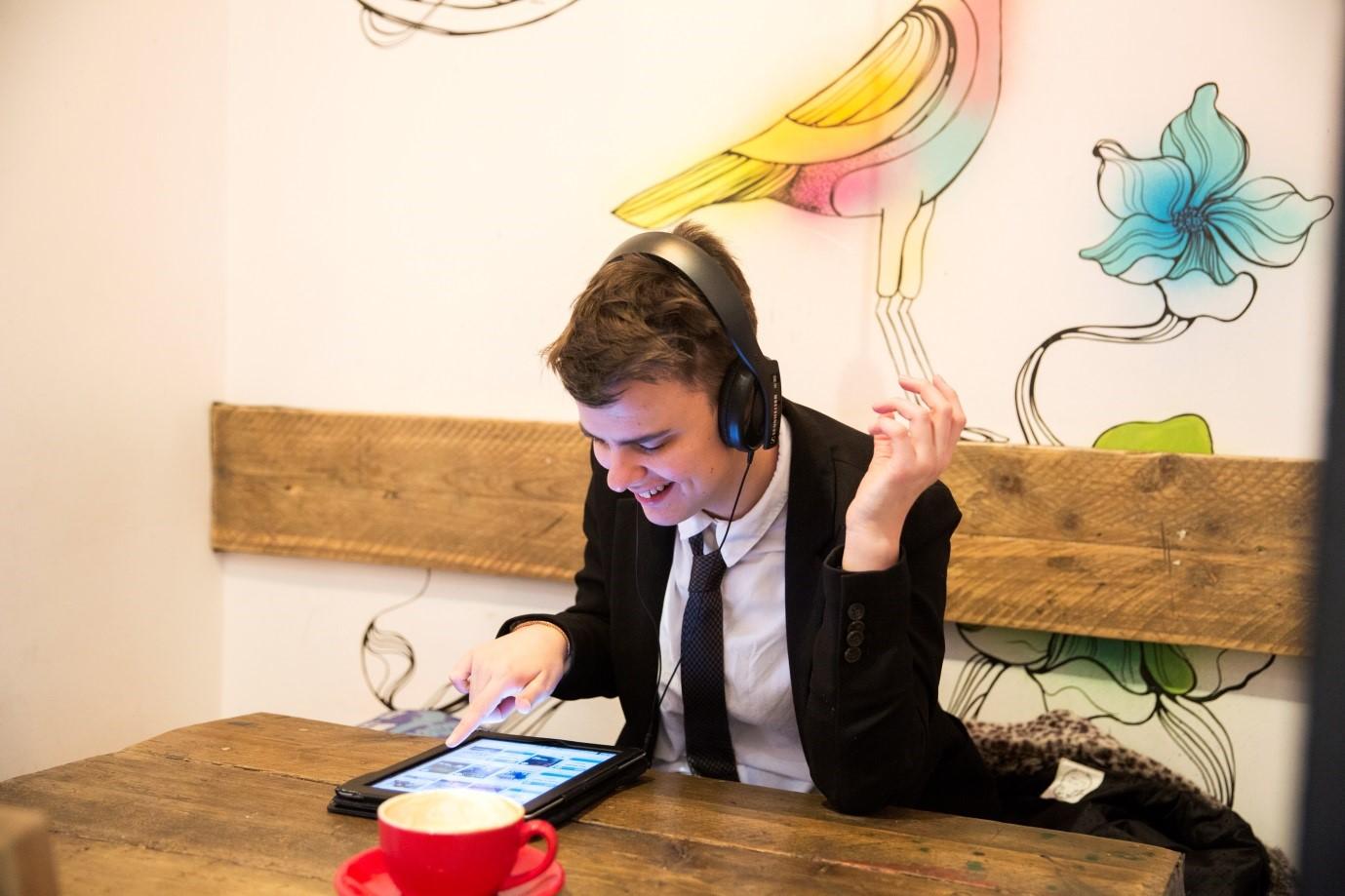 a laughing school boy in uniform, sat at a table with coffee cup using his iPad wearing headphones. There a decorative mural on the wall behind him.